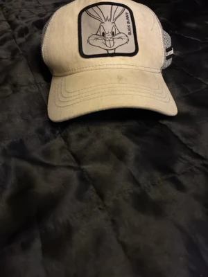 Beige Bugs Bunny keps - Säljer en beige keps med svartvit Bugs Bunny-patch framtill. Kepsen har mesh på sidorna och bak för extra ventilation och justerbar passform. Perfekt för dig som gillar Looney Tunes och vill ha en unik accessoar.