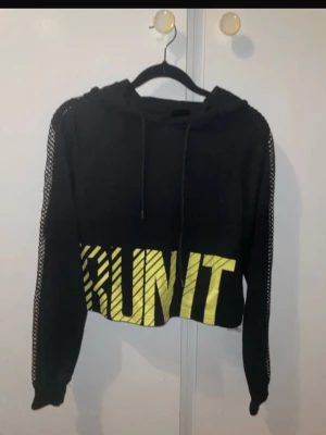 Svart croppad hoodie med meshärmar - Cool svart croppad hoodie med huva och meshdetaljer längs ärmarna. Stora gula bokstäver med texten 'RUN IT' framtill ger en sportig vibe. Perfekt för dig som gillar streetstyle och vill sticka ut.