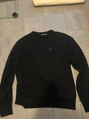 Svart sweatshirt från Acne Studios - En stilren svart sweatshirt från Acne Studios med rund halsringning och diskret logopatch på bröstet. Tröjan har långa ärmar och ribbade muddar vid ärmslut och nederkant. Perfekt för en clean och modern look.