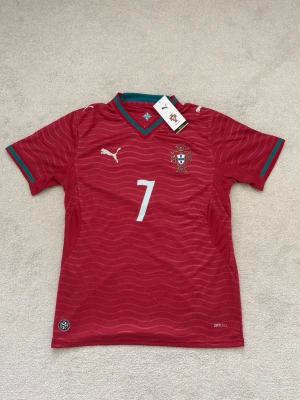 Portugal Ronaldo matchtröja Puma - Snygg röd Portugal matchtröja från Puma med nummer 7 och Ronaldo på ryggen. Tröjan har gröna och röda detaljer vid krage och ärmslut, samt vågigt mönster i tyget. Tillverkad i lätt och ventilerande material med DryCell-teknologi. Perfekt för fotbollsträning eller support.