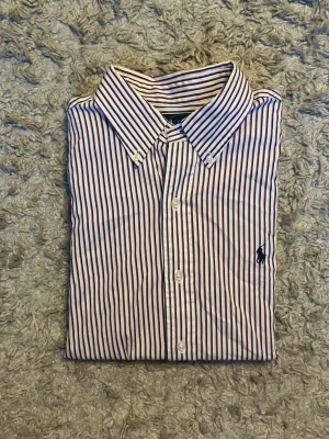 Randig skjorta från Ralph Lauren - Klassisk skjorta från Ralph Lauren med smala blå och vita ränder. Skjortan har lång ärm, knappar framtill och ikoniska broderade loggan på bröstet. Tillverkad i mjuk bomull och har en tidlös button-down krage.