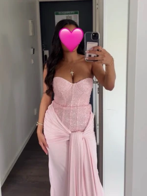 Carina Maxi Dress - Blush från Babyboo - Elegant ljusrosa långklänning med paljettprydd överdel och draperad kjol. Klänningen är axelbandslös och har en snygg omlott-detalj framtill. Perfekt för speciella tillfällen. (Klänningen är lite kort för att vi sydde upp den lite)