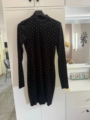 Svart prickig långärmad klänning - Snygg svart kortklänning med vita prickar och lång ärm. Klänningen har en hög krage och figurnära passform, perfekt för dig som gillar en stilren look med lite edge. Materialet känns mjukt och stretchigt, vilket gör den bekväm att bära.