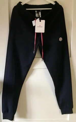 Svarta mjukisbyxor från Moncler - Snygga svarta mjukisbyxor från Moncler med elastisk midja och dragsko i rött, vitt och blått. Byxorna har Moncler-logga på vänster ben och ribbade muddar vid bensluten. Perfekta för en chill och trendig look.