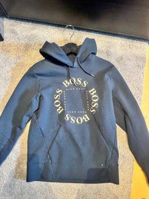 Hugo boss hoodie  - Snygg blå hoodie från Hugo Boss med BOSS-logga i cirkel på bröstet. Klassisk huva med dragsko och känguruficka framtill. Tillverkad i mjukt bomullsmaterial som är skönt att bära. Perfekt för en avslappnad och trendig look.