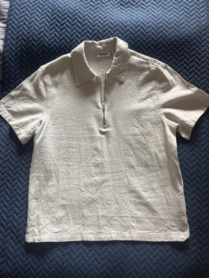 Beige kortärmad topp från Weekday - Säljer en beige linné skjorta från Weekday med korta ärmar och krage. Skjortan har en clean look med half zip-dragkedja framtill. Perfekt för dig som gillar minimalistisk stil.