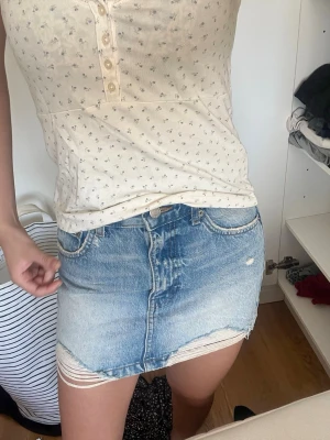 Blå jeanskjol med slitna detaljer - Trendig kort jeanskjol i ljusblå denim med slitna kanter och rå fåll. Klassisk femficksmodell med dragkedja och knapp framtill. Perfekt för en avslappnad och cool stil.