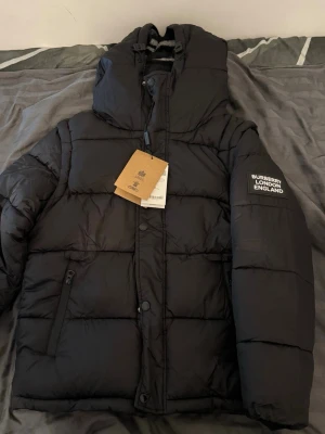 Svart pufferjacka från Burberry - Pris kan diskuteras vid snabb affär. Kan bli till en väst. Snygg svart pufferjacka från Burberry med huva och dragkedja framtill. Jackan har en patch med texten 'BURBERRY LONDON ENGLAND' på ärmen och innerfoder i klassiskt rutmönster. Perfekt för kalla vinterdagar och riktigt trendig stil.
