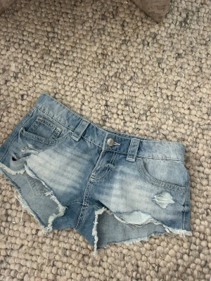 Ljusblå jeansshorts med slitningar - Säljer ett par ljusblå jeansshorts med råa kanter och slitna detaljer framtill. Shortsen har klassisk femficksdesign, knappgylf och fransiga benslut för en avslappnad look. Perfekta för varma dagar och enkel att matcha.