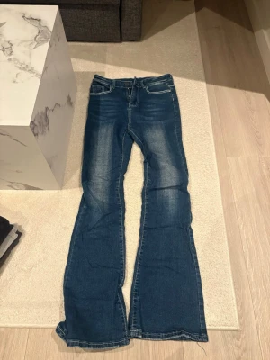 Blå bootcut jeans med broderade fickor - Säljer ett par blå bootcut jeans med snygga broderade detaljer på bakfickorna och vita kontrastsömmar. Jeansen har klassisk femficksmodell och utsvängda ben. Perfekta för dig som gillar en retroinspirerad look. De har ett pyte lotter märka i form av en penna men det syns verkligen inte. 