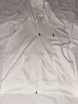 Vit hoodie från Polo Ralph Lauren - Snygg vit hoodie från Polo Ralph Lauren med dragkedja framtill och klassisk logga broderad på bröstet. Hoodien har huva med dragsko och två stora fickor framtill. Tillverkad i mjukt bomullsmaterial som känns skönt mot huden.