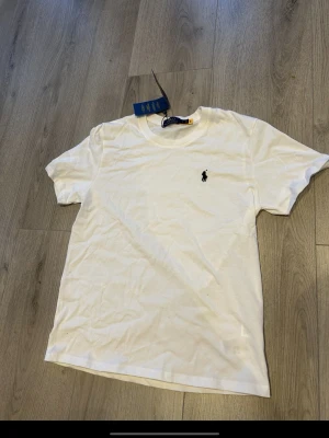 Vit t-shirt från Ralph Lauren - Klassisk vit t-shirt från Ralph Lauren med den ikoniska svarta polospelaren broderad på bröstet. T-shirten har rund hals, korta ärmar och är gjord i mjuk bomull för en skön känsla. Perfekt basplagg till garderoben. Står M men passar Som S