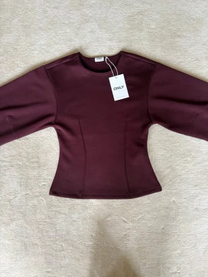 Figursydd vinröd topp - Trendig vinröd topp från ONLY med markerad midja och vida, långa ärmar. Toppen har en clean look utan mönster och är tillverkad i ett stretchigt material som ger en snygg passform. Perfekt för dig som vill ha en stilren och modern vibe. Helt ny, nypris 449kr (pris kan diskuteras)💕