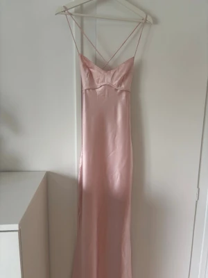 Ljusrosa långklänning med smala band - Elegant ljusrosa långklänning i satin med tunna axelband som korsas i ryggen och snörning baktill. Klänningen har en figurnära passform och en mjukt skimrande yta som ger en lyxig känsla. 140cm lång utan axelband. Använd en kväll. Säljer för att den inte kommer användas igen. Köpt för 1400kr med frakt. Skriv för frågor 