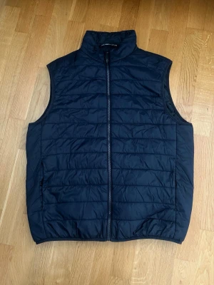 Mörkblå dunväst från Zara | Storlek M - Stilren svart dunväst från Zara i storlek M med hög krage och dragkedja framtill. Västen har ett quiltat mönster och två sidofickor med dragkedja. Perfekt att slänga över en hoodie eller tröja för extra värme. Lätt och smidig att bära men en personlig favorit till sommaren. Säljer då den är för stor.