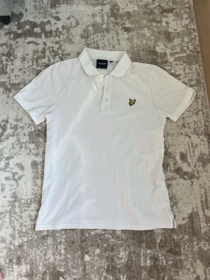 Vit pikétröja från Lyle & Scott - Klassisk vit pikétröja från Lyle & Scott med korta ärmar och gul logga på bröstet. Tröjan har krage och knappar framtill, perfekt för en clean och stilren look.