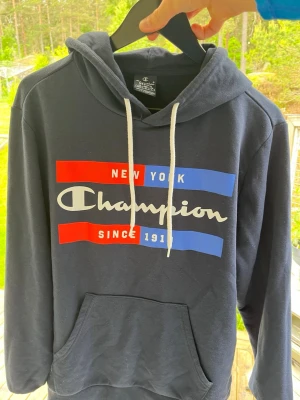 Mörkblå Champion hoodie med tryck - Snygg mörkblå hoodie från Champion med stor logga och texten 'New York' och 'Since 1919' i rött, vitt och blått framtill. Klassisk känguruficka och vita dragsnören i huvan. Perfekt för en avslappnad streetwear-look. Pris kan diskuteras 