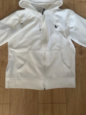 Vit hoodie från Polo Ralph Lauren - Snygg vit hoodie från Polo Ralph Lauren med dragkedja framtill och klassisk broderad logga på bröstet. Hoodien har kängurufickor och en stor huva. Tillverkad i mjuk bomull för skön känsla och stilren look.