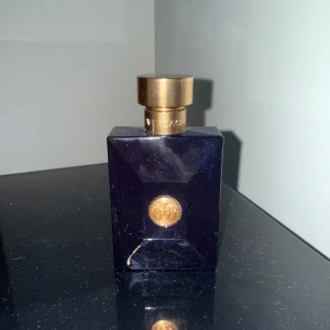 Mörkblå parfymflaska med gulddetaljer - En versace  mörkblå parfymflaska med en fyrkantig form och guldfärgad kork. Flaskan har en gyllene emblem på framsidan som ger en lyxig känsla. Perfekt för den som söker en stilren och exklusiv doftupplevelse. Den är inte helt full