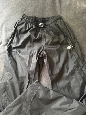 Svarta mjukisbyxor från Nike - Snygga svarta mjukisbyxor från Nike med klassisk logga broderad på vänster ben. Byxorna har resår i midjan med dragsko och är tillverkade i ett lätt, glansigt material som ger en sportig vibe. Perfekta för chill eller träning.