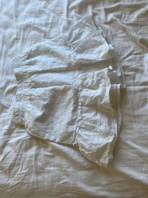 Vit volangkjol med broderade detaljer - Söt vit kortkjol med volang och broderade blommor. Kjolen har resår i midjan och är gjord i ett lätt bomullstyg, perfekt för varma dagar. Den romantiska stilen och de små broderierna ger en somrig känsla.