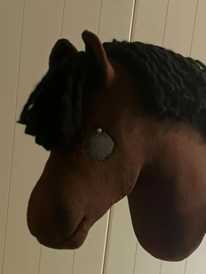 Brun käpphäst med svart man - Säljer en handgjord käpphäst i mjukt brunt tyg med svart garnman och sydda detaljer. Perfekt för lekar och hobbyridning. Hästens ögon är sydda i svart tyg och den har en klassisk hästformad design.