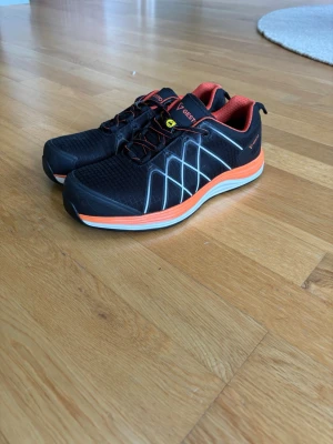 Svarta sneakers från Gesto med orange detaljer - Snygga svarta stål sneakers från Gesto med orange och vita detaljer. Skorna har meshmaterial på ovansidan, reflexliknande mönster och orange foder. Låg modell med klassiska snörningar och bekväm sula. Perfekt för dig som har ett praktiskt jobb och behöver nya ståldojjor. Skorna är väldigt bra skick och använda kanske 5 gånger. Storlek 42