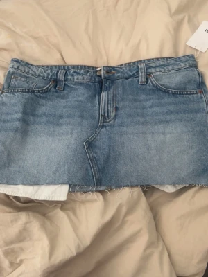 Blå jeanskjol med rå kant - Säljer en kort blå jeanskjol med rå, fransig kant och klassiska fem fickor. Kjolen har bälteshällor och knappgylf framtill. Perfekt för dig som gillar en avslappnad och trendig denimlook. Storlek 44 och i nyskick med prislapp på