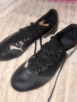 Svarta Puma fotbollsskor med dobbar - Säljer ett par svarta fotbollsskor från Puma med vita detaljer och klassisk logga på sidan. Skorna har snörning och slitstarka dobbar för bra grepp på planen. Ovandelen är i syntetmaterial som ger en tight passform och bra bollkontroll. Perfekta för dig som vill ta spelet till nästa nivå.