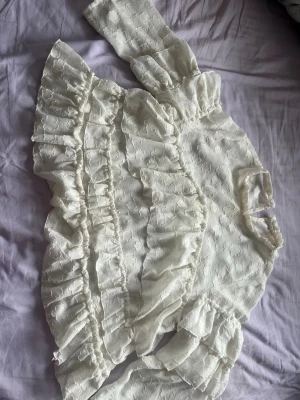 Vit volangblus med spetsdetaljer - Supersöt vit blus med volanger och blommig spets. Blusen har puffiga långärmar och hög hals med volangkant. Materialet är tunt och genombrutet med blommönster, vilket ger en romantisk och trendig look. Perfekt att styla med jeans eller kjol.
