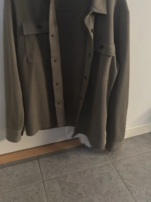 Olivgrön overshirt från Selected - Stilren olivgrön overshirt från Selected med klassisk krage, tryckknappar och två stora bröstfickor. Overshirten har en rak passform och är tillverkad i ett kraftigare tyg som ger en snygg och avslappnad look.knappt använd 