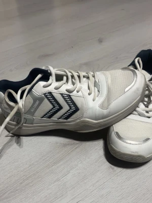 Vita inomhusskor från Hummel - Säljer ett par vita inomhusskor från Hummel med mörkblå detaljer och klassiska pilar på sidan. Skorna har snörning, meshpaneler för ventilation och slitstark sula. Perfekta för sporter som handboll eller innebandy. Endast använda 2 gånger. Nypris 680kr