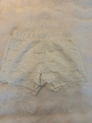 Vita spetsdetaljerade shorts - Säljer ett par vita shorts med bred resår i midjan och flera lager av spetsvolanger. Shortsen har en romantisk vibe med blommiga spetsmönster och mjukt tyg som känns skönt mot huden. Perfekta för varma dagar när du vill ha något både sött och bekvämt. Stolek 158/164 (xs)