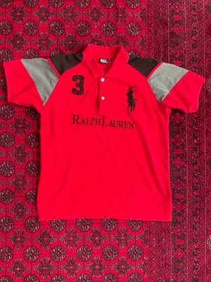 Ralph Lauren Pike - Säljer en röd Polo skjorta från Ralph Lauren med svarta och grå detaljer på axlarna. Skjortan har klassisk krage, knappar framtill och broderad logga samt siffran 3 på bröstet. Materialet är mjuk bomull och passformen är normal.