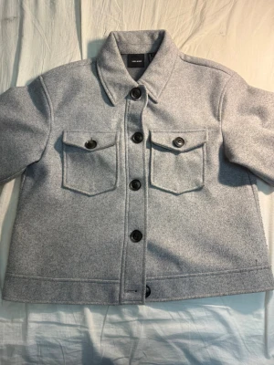 Ljusgrå overshirt från Weekday - Stilren ljusgrå overshirt från veramoda med klassisk krage och stora svarta knappar. Två bröstfickor med lock och knapp framtill. Overshirten är gjord i ett mjukt, filtliknande material som ger en modern och avslappnad look.