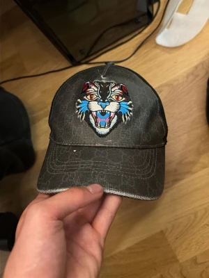 Svart keps med tigerbrodyr - Säljer en svart keps med ett stort broderat tigerhuvud framtill i färgerna blått, rött och vitt. Kepsen har böjd skärm och justerbar rem bak. Materialet ser ut att vara bomullsmix med diskret mönster i tyget. Perfekt för dig som vill sticka ut med en cool accessoar.