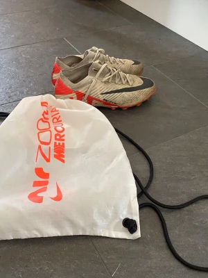 Nike Air Zoom Mercurial fotbollsskor - Säljer ett par Nike Air Zoom Mercurial fotbollsskor i vit färg med röda och svarta detaljer. Skorna har snörning, låg profil och mönstrad sula för bra grepp på planen. Tillverkade i lätt syntetmaterial för smidighet och snabbhet. Perfekta för dig som vill ha stil och prestanda på matchen.