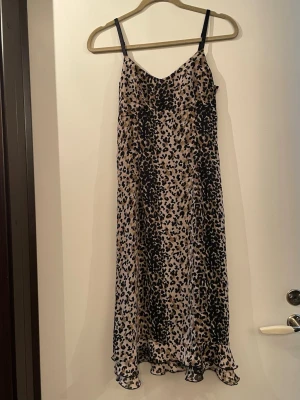 Leopardmönstrad elegant klänning  - Säljer en snygg midiklänning med leopardmönster i beige och svart. Klänningen har smala axelband och v-ringad halsringning. Materialet känns lätt och svalt, perfekt för varma dagar. Volangdetalj längst ner ger extra rörelse till looken.