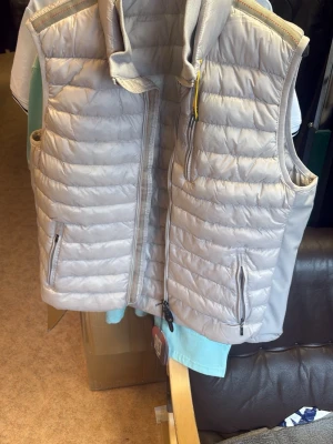 Ljusgrå dunväst från Parajumpers - Säljer en snygg ljusgrå dunväst från Parajumpers, modell 1099. Västen har quiltad design, hög krage och två dragkedjeförsedda fickor framtill. Materialet är lätt och isolerande, perfekt för lager-på-lager. Färgen är 'stone' och den har coola detaljer som gul dragkedja vid kragen.