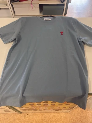 Blå t-shirt från AMI Paris - Snygg blå t-shirt från AMI Paris med klassisk passform och korta ärmar. På bröstet finns ett rött broderat hjärta med A-logga. Tillverkad i mjuk bomull som känns skön mot huden. Perfekt för en stilren och avslappnad look.