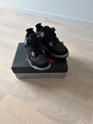 Svarta Air Jordan 4 Retro sneakers - Säljer ett par svarta Air Jordan 4 Retro sneakers med grå detaljer och meshpaneler på sidorna. Skorna har en chunky sula med synlig Air-enhet och coola plastdetaljer vid snörningen. Perfekta för dig som gillar streetwear och klassisk sneakerstil.