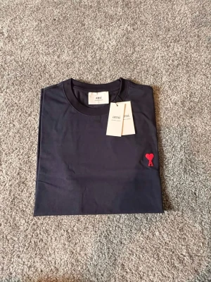 Ami Paris T-shirt mörkblå Ny-oanvänd (M) - AMI Paris T-shirt – Mörkblå (Storlek M) Säljer en helt ny och oanvänd AMI Paris T-shirt i storlek M. Stilren och modern design i mörkblå färg med AMI Paris ikoniska logga som ger en exklusiv och minimalistisk look. Tillverkad i hög kvalitet med mjukt och bekvämt material – perfekt för både vardag och streetwear-stil. 🔹 Märke: AMI Paris 🔹 Modell: T-shirt 🔹 Färg: Mörkblå 🔹 Storlek: M 🔹 Skick: Helt ny / oanvänd 🔹 Passform: Klassisk Nypris är högt – perfekt möjlighet att göra ett fynd