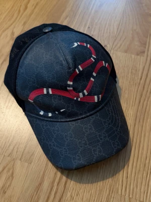 Gucci keps med orm  - Snygg svart keps från Gucci med ikoniskt GG-mönster och en röd-vit orm broderad på framsidan. Baksidan har mesh-tyg för extra ventilation och justerbar rem. Perfekt för dig som vill sticka ut med en exklusiv accessoar.
