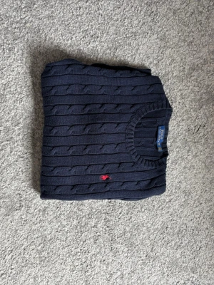 Polo Ralph Lauren - Kabelstickad mörkblå tröja från Polo Ralph Lauren med rund halsringning och röd broderad logga på bröstet. Tröjan har långa ärmar och ribbade muddar vid ärmslut och nederkant. Perfekt för en klassisk och stilren look.