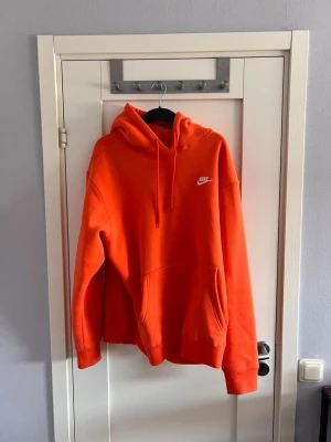 Orange hoodie från Nike med huva - Snygg orange hoodie från Nike med klassisk logga på bröstet. Hoodien har stor känguruficka framtill, justerbar dragsko i huvan och ribbade muddar vid ärmar och nederkant. Perfekt för en avslappnad och sportig stil.