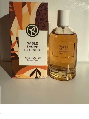 Sable Fauve Eau de Parfum Yves Rocher - Sable Fauve Eau de Parfum från Yves Rocher i en elegant rund glasflaska med vit kork. Doften kommer i en snygg förpackning med abstrakt mönster i brunt, orange och beige. Flaskan rymmer 100 ml och har en varm bärnstensfärgad vätska.