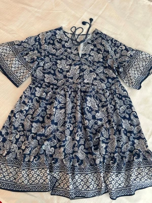 Blåvit kortklänning med paisleymönster M, längd 86 cm - Kortklänning i blått och vitt med paisley- och bårdmönster. Klänningen har v-ringning med snörning, vida trekvartsärmar och avslappnad passform. Perfekt för sommarens varma dagar och har en bohemisk vibe.