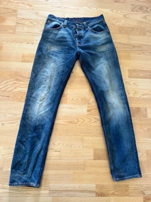 Blå vintage jeans från Nudie Jeans - Säljer ett par klassiska blå jeans från Nudie Jeans med snygg slitning och raka ben. Jeansen har fem fickor, orange sömmar och är gjorda i kraftig denim. Perfekta för dig som gillar en avslappnad och tidlös stil.