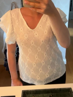 Vit blus med volangärm och spetsdetaljer - Söt vit blus med v-ringning och korta volangärmar. Blusen har ett broderat spetsmönster i hela tyget. Den är från Vila och är köpt för ca 200kr. Enbart testad