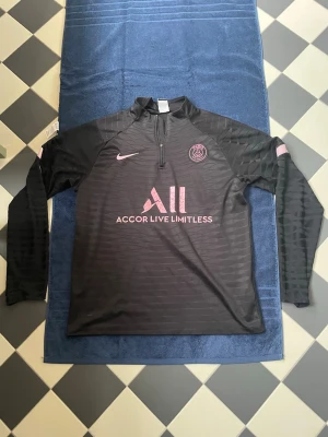 Svart PSG träningströja från Nike - Snygg svart långärmad träningströja från Nike med PSG-logga och rosa detaljer. Halvlång dragkedja framtill och trycket 'Accor Live Limitless' på bröstet. Tillverkad i ett lätt och funktionellt material som passar perfekt till träning. Tecken på användning men den fungerar ändå. 